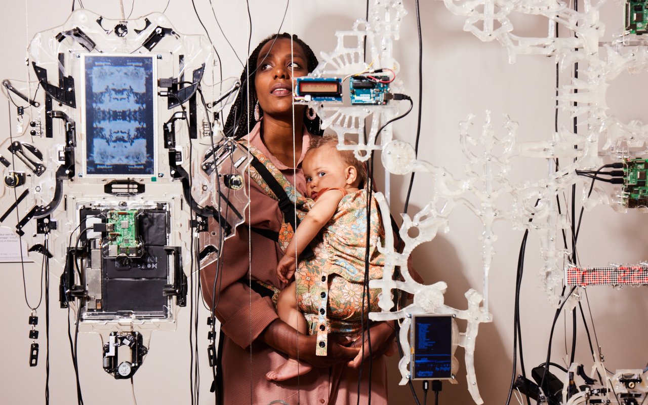 Jonge vrouw met baby in draagzak staat achter een opgehangen kunstwerk waar je haar doorheen ziet. De baby kijkt in de camera.