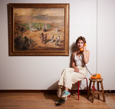 Jonge vrouw zit op een stoeltje naast een kunstwerk. Ze heeft een ouderwetse telefoon met draad in haar hand.