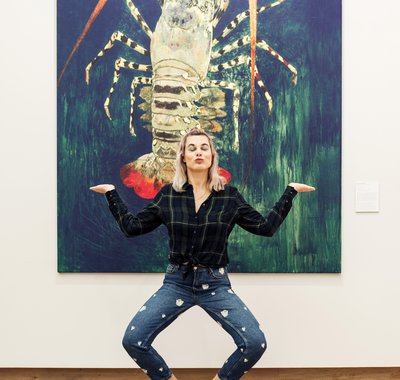 Vrouw staat in een pose voor het grote en kleurrijke schilderij van een kreeft.