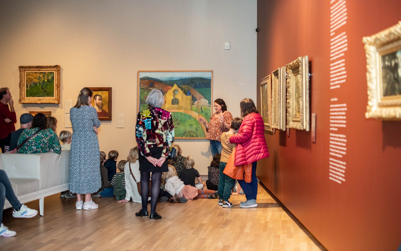 Een groep mensen staat en zit in een museumzaal. Ze krijgen uitleg van een vrouw.