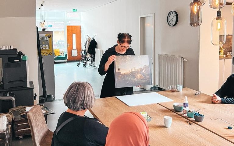 Iemand houdt een schilderij op en vertelt daarover aan een groep mensen die aan tafel zit.