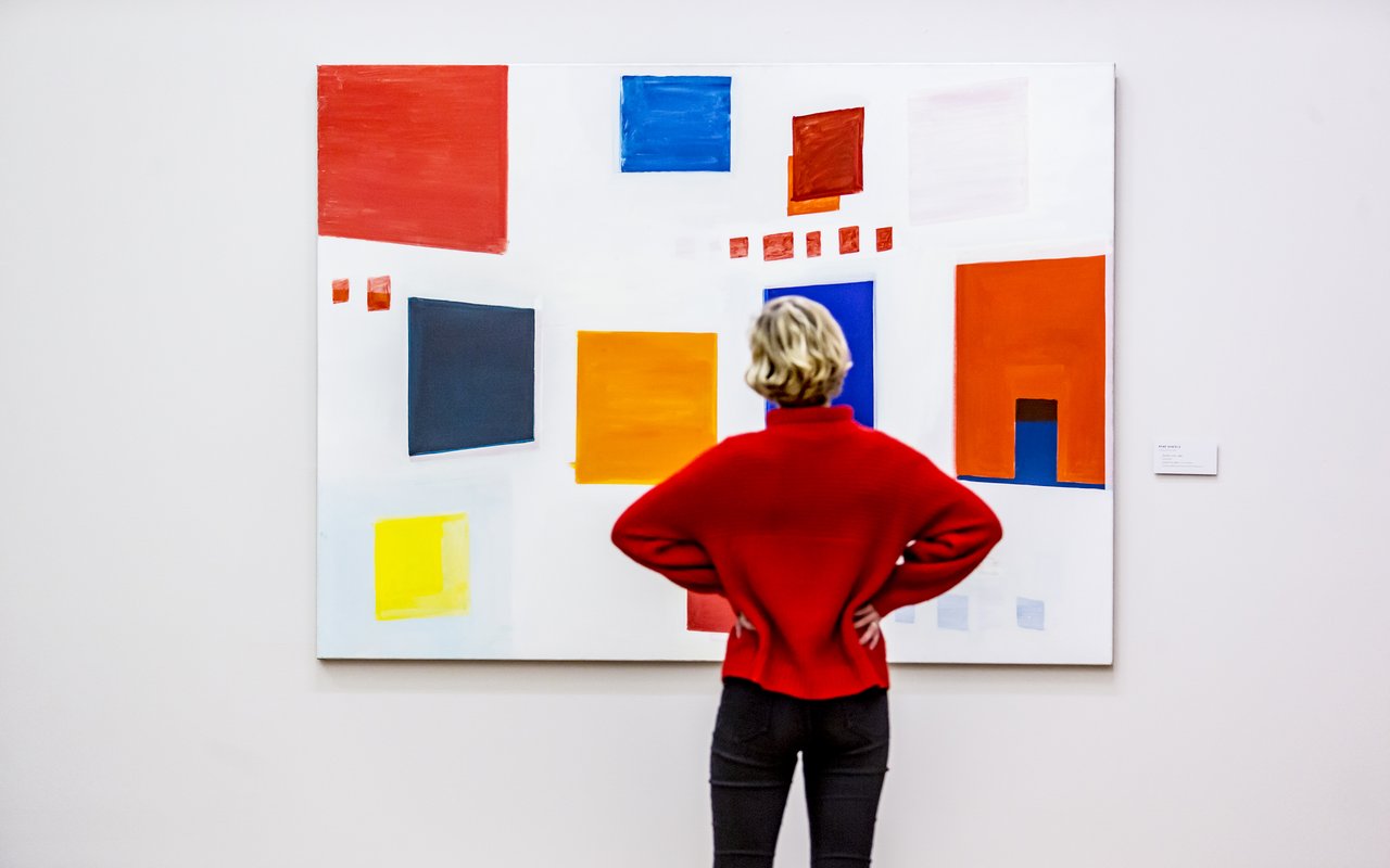 Vrouw in rode trui staat voor een schilderij met gekleurde vlakken.
