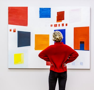Vrouw in rode trui staat voor een schilderij met gekleurde vlakken.