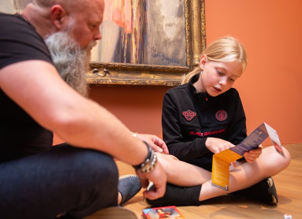 Een vader en zijn dochter lezen samen de instructie van het Museumdobbelspel. Een vader en zijn dochter lezen samen de instructie van het Museumdobbelspel.