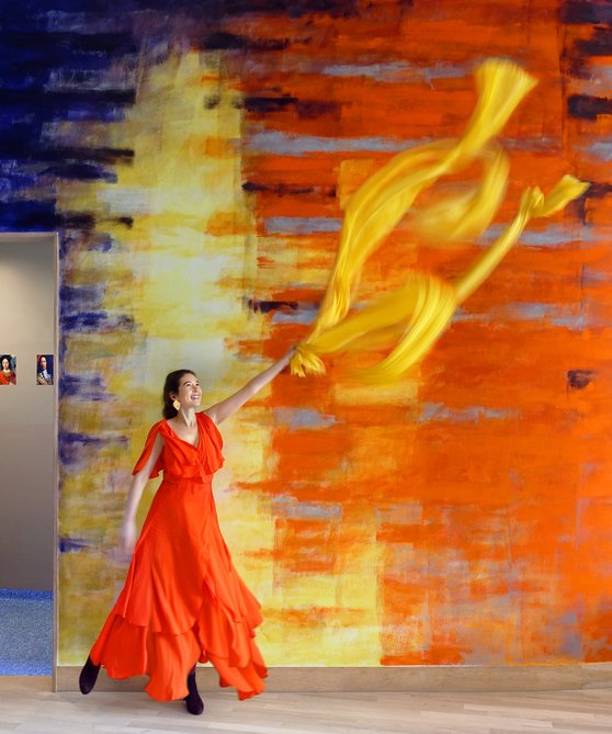 Vrouw in oranje jurk zwaait met gele sjaal. Ze staat voor het geel/oranje deel van de muurschildering.