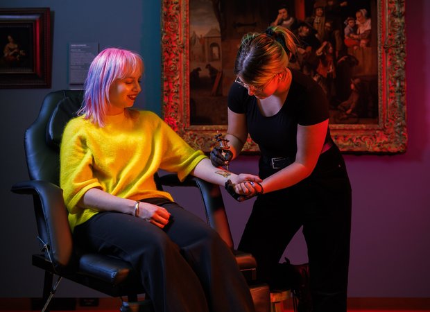 Een jonge vrouw zit in een stoel in een zaal van Dordrechts Museum en krijgt een neptattoo op haar gebracht door een andere jonge vrouw.