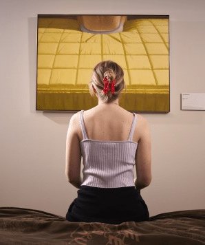 Een jonge vrouw, vanaf de rug gefotografeerd, zit op een bankje voor het schilderij Bed van Jan Beutener.