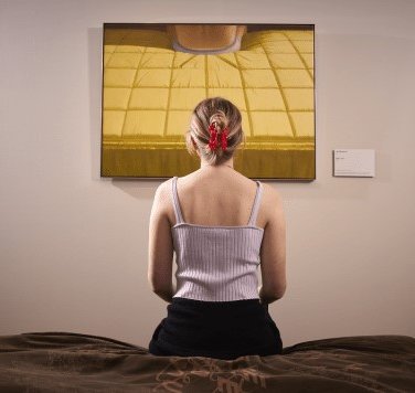 Een jonge vrouw, vanaf de rug gefotografeerd, zit op een bankje voor het schilderij Bed van Jan Beutener.