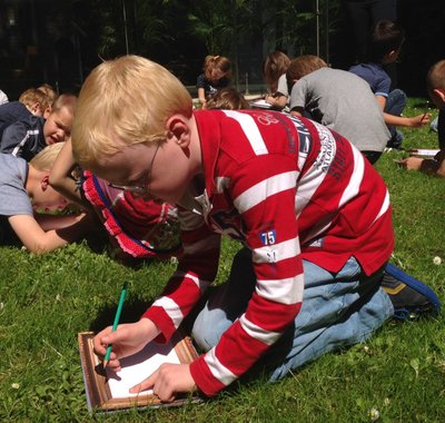 In de tuin zitten kinderen op hun knieën te tekenen. Vooraan een jongetje in een rood wit gestreepte trui.