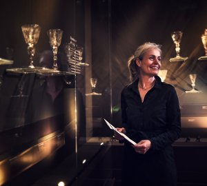 Lachende vrouw met folder in haar hand staat bij de glascollectie.