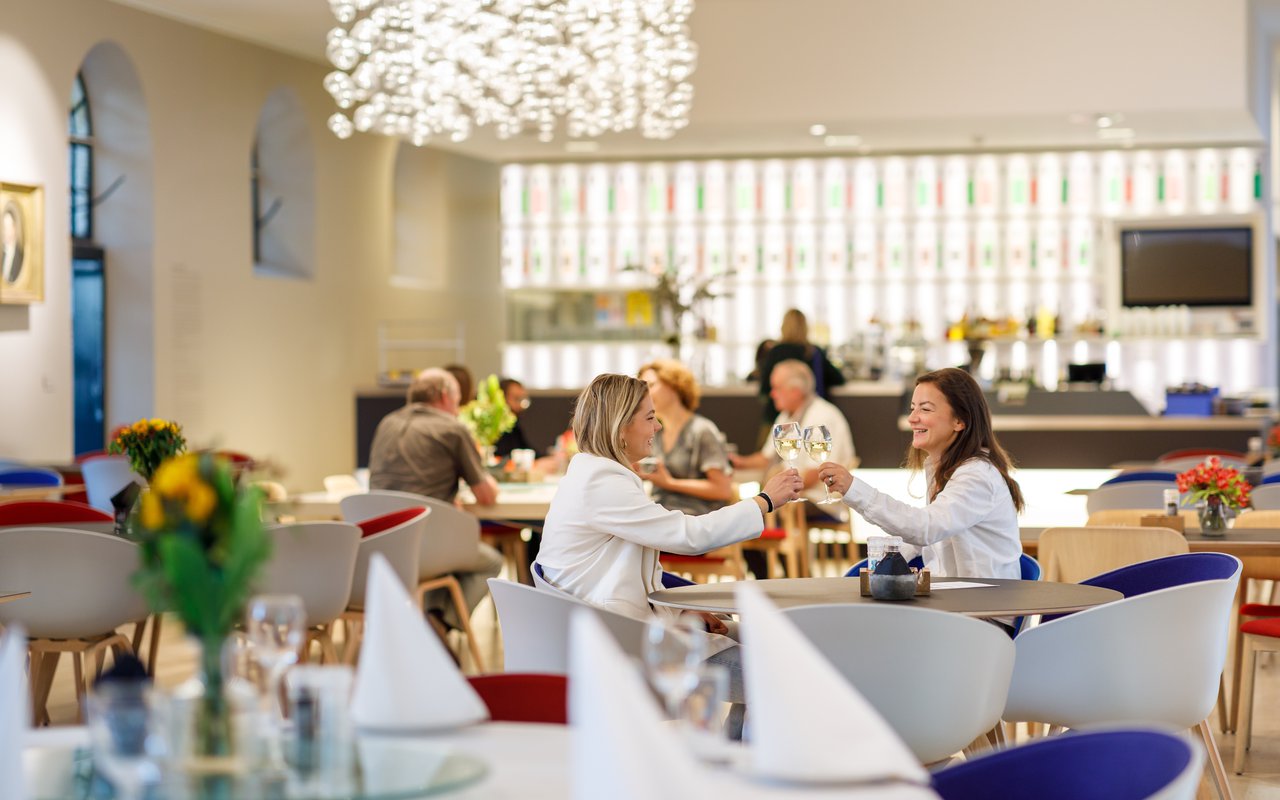 In het Museumrestaurant zitten mensen aan tafeltjes Twee vrouwen proosten.